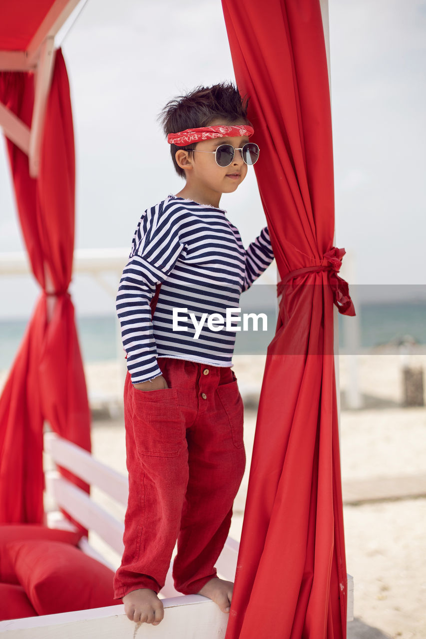 Stylish baby boy in a striped t-shirt and sunglasses stands at the red beach sunbed