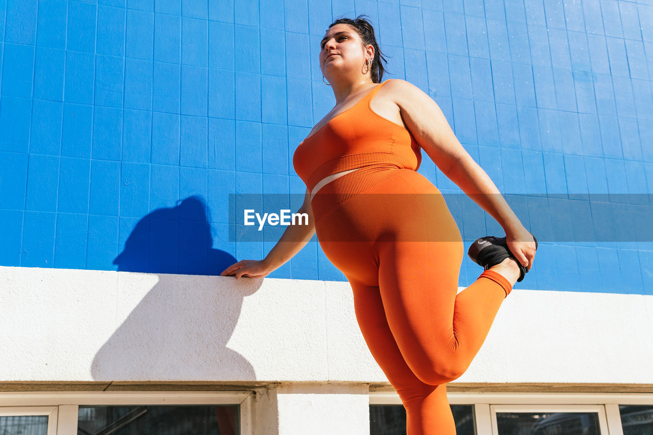 From below plump female athlete in sportswear exercising on tiled walkway in sunny town