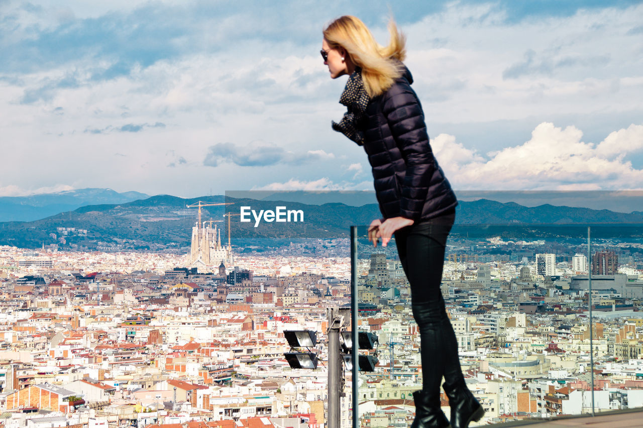 Young woman wearing warm clothing while standing on balcony against cityscape