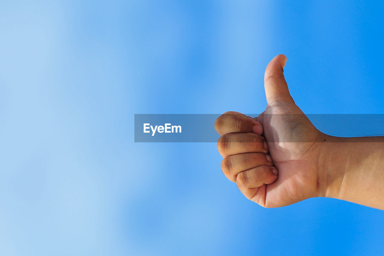 Low angle view of hand showing thumbs up sign against clear blue sky