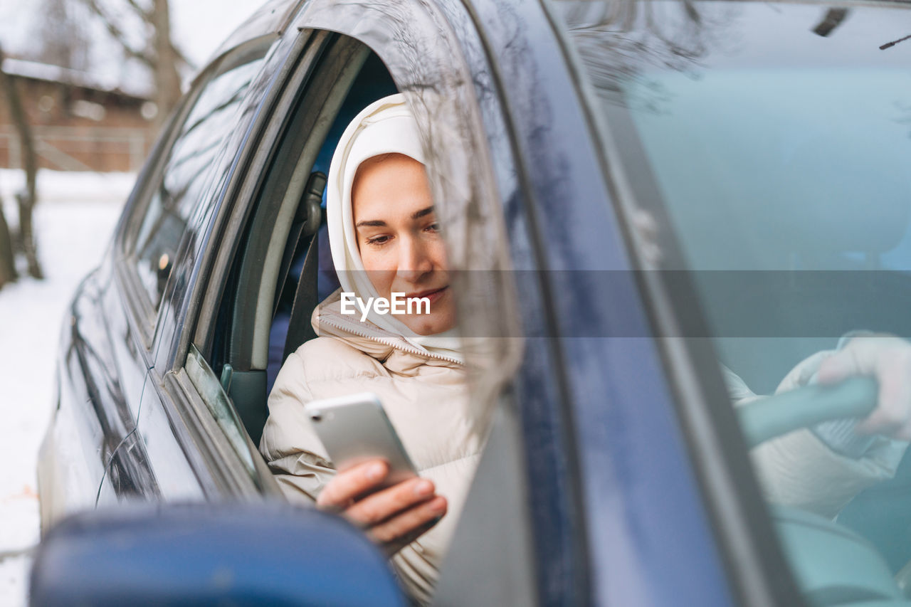Beautiful smiling young muslim woman in headscarf  using mobile in right-hand-drive car