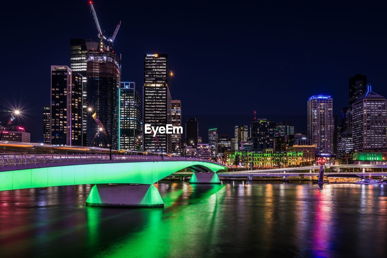 Illuminated bridge over river by buildings against sky at night