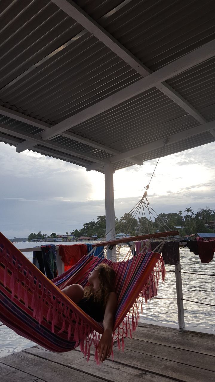 Woman lying on hammock in gazebo over sea against sky during sunset
