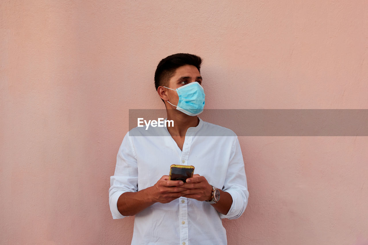 Young latin man with mask is looking his phone on pink background