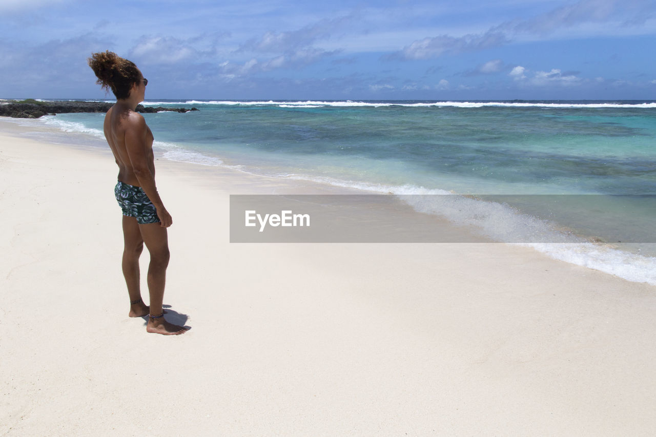 Tanned man at beach, with curly hair and bun, staring at the blue sea
