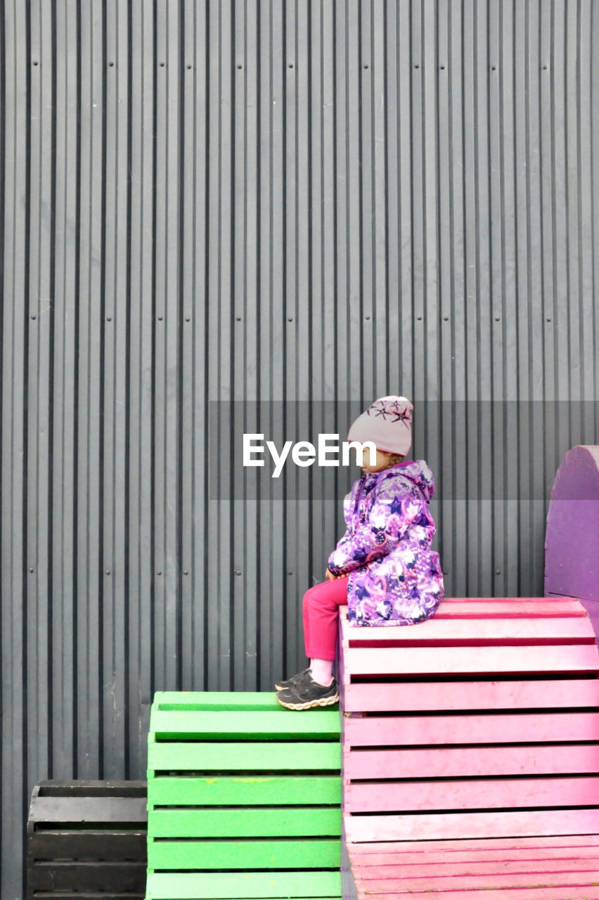 Side view of girl in warm clothing looking away while sitting on bench against wall