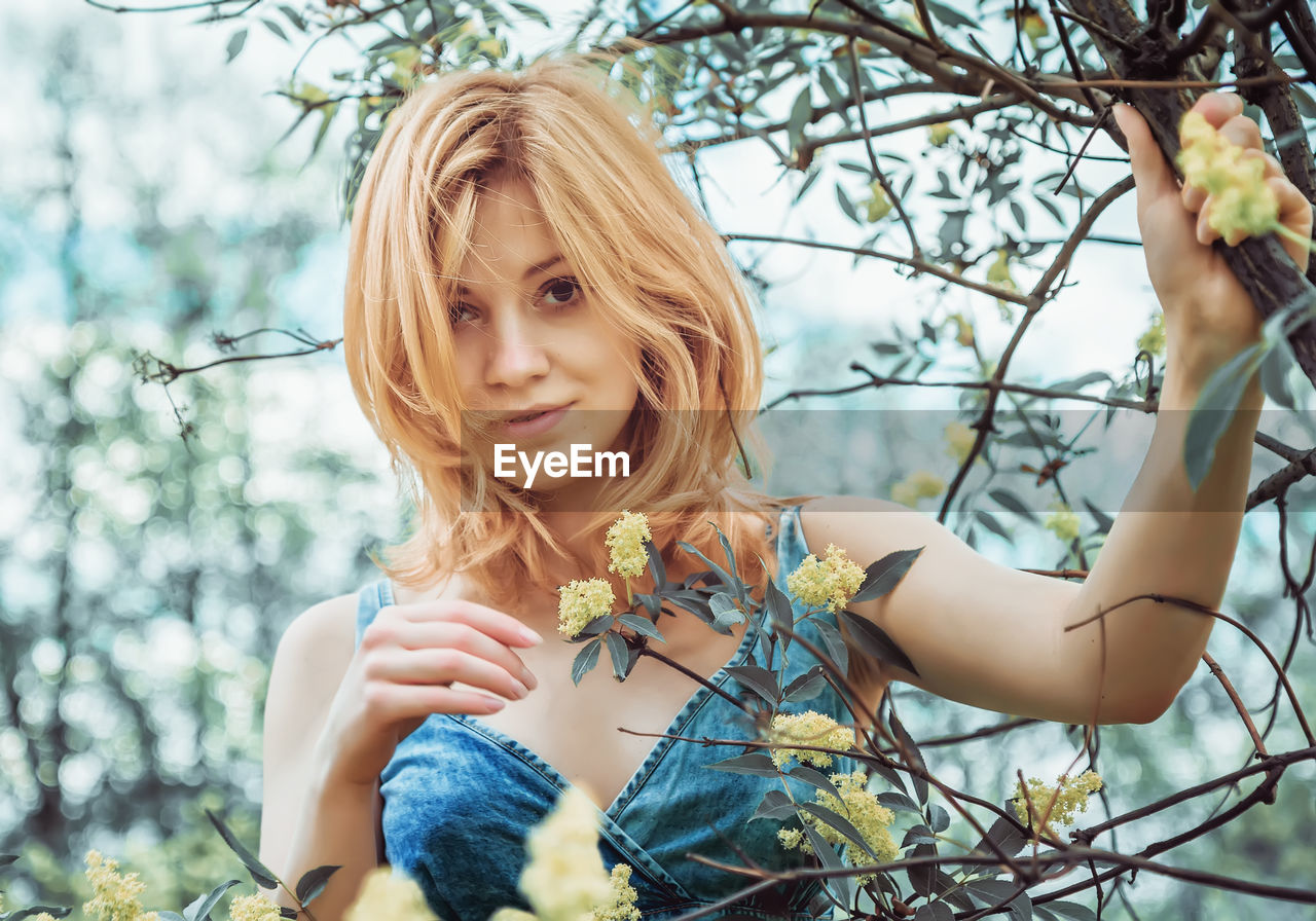 Young blonde woman in a denim top stands among flowering branches in nature and looks at the camera.