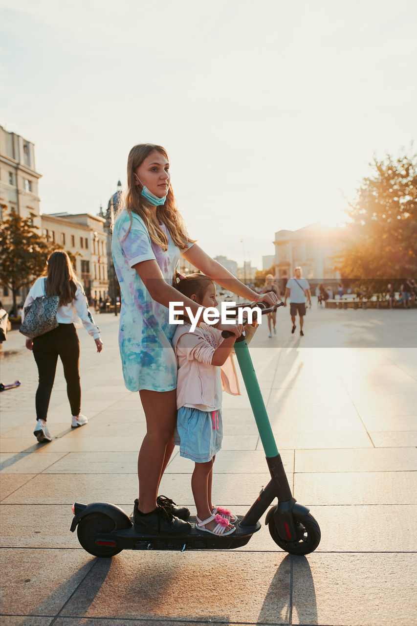 Teenager girl and her sister preschooler riding an electric scooter in the city center