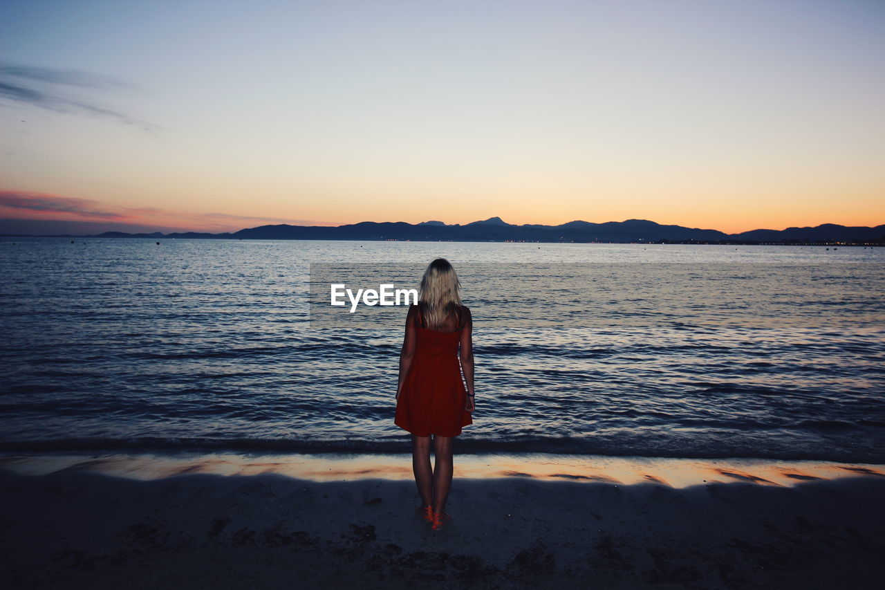 WOMAN STANDING AT BEACH AGAINST SKY DURING SUNSET