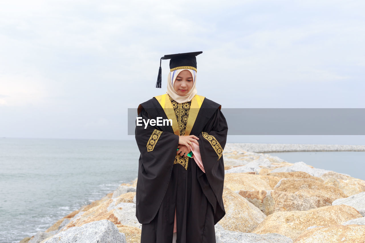 Young woman in graduation gown standing at beach against cloudy sky