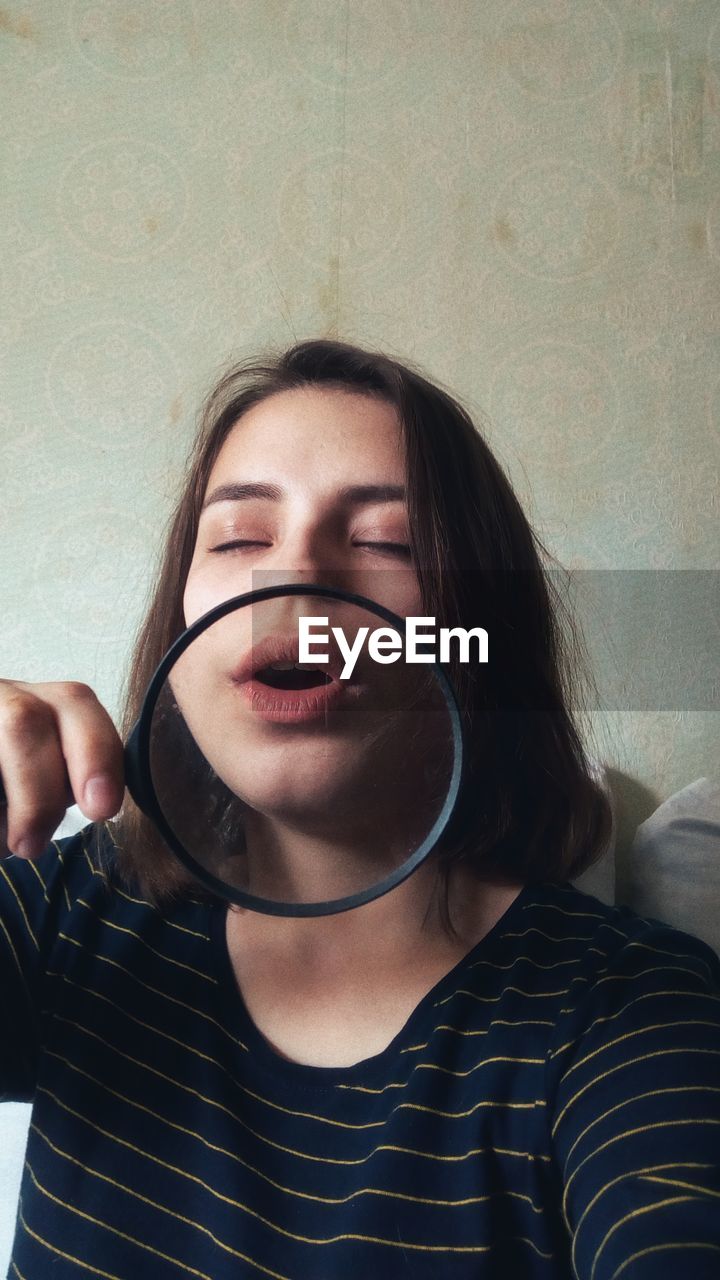 Close-up of young woman holding magnifying glass against wall