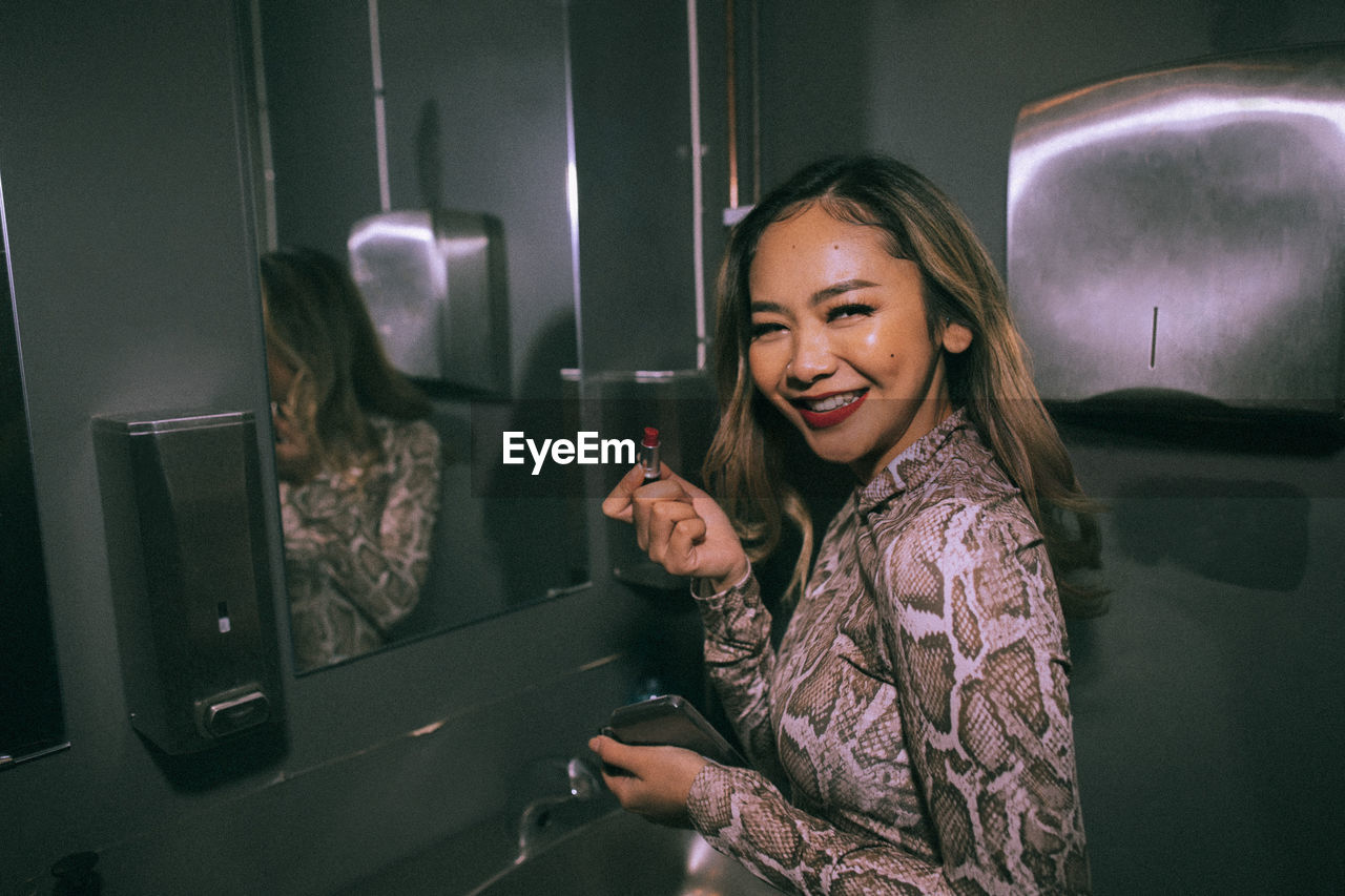 Portrait of smiling young woman applying lipstick in bathroom at nightclub