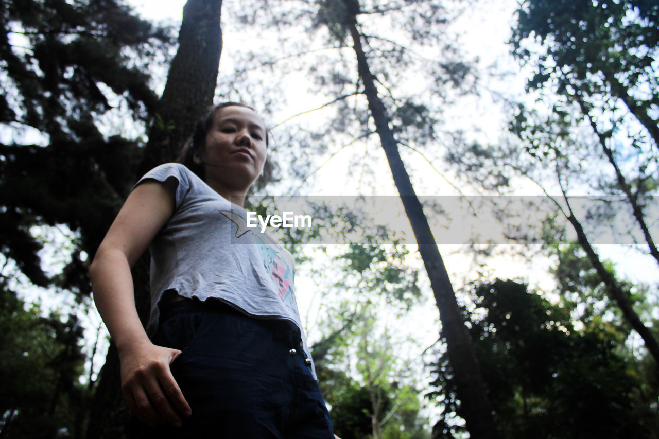 Low angle portrait of woman standing against tree in forest