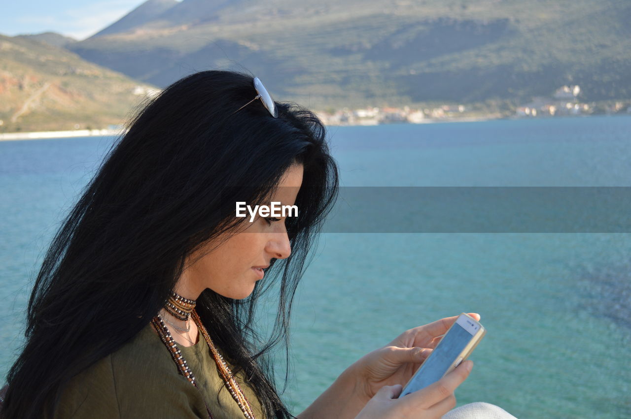 Side view of young woman using phone against sea