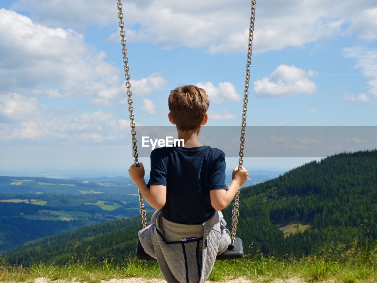 Rear view of boy swinging by landscape against cloudy sky