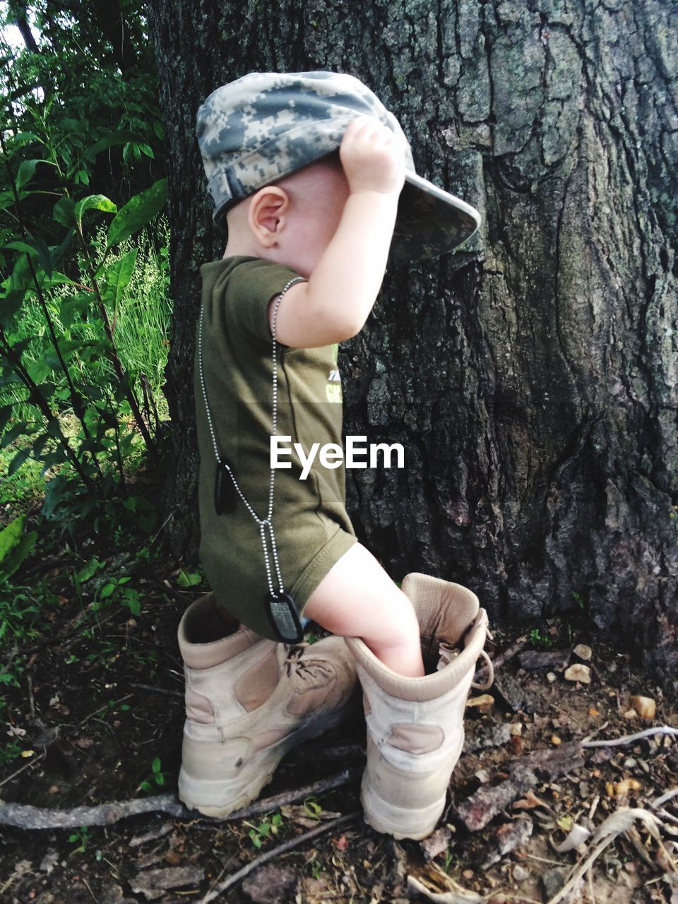 Cute toddler wearing military boots and cap against tree