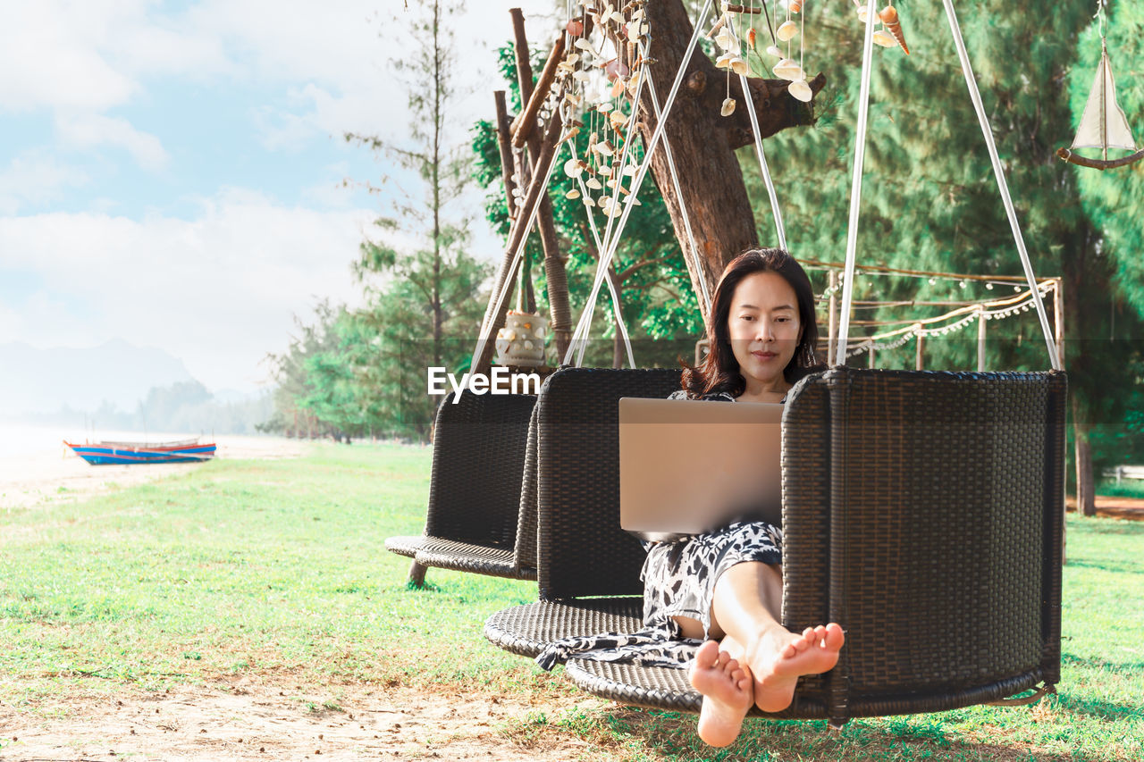 Beautiful woman working laptop while sitting in hammock on the beach .