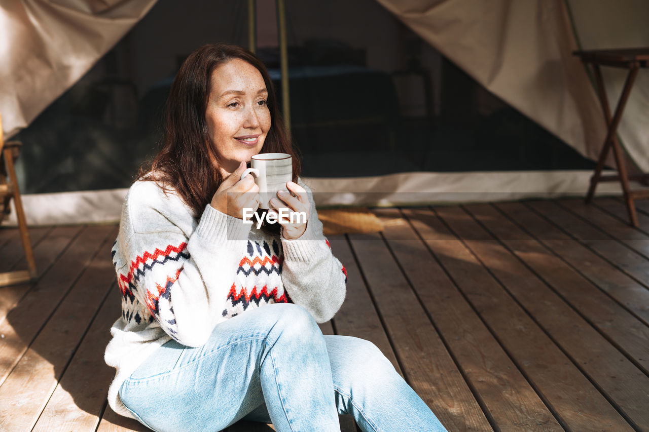 Brunette woman in nordic sweater drinking tea and relaxing in glamping in nature