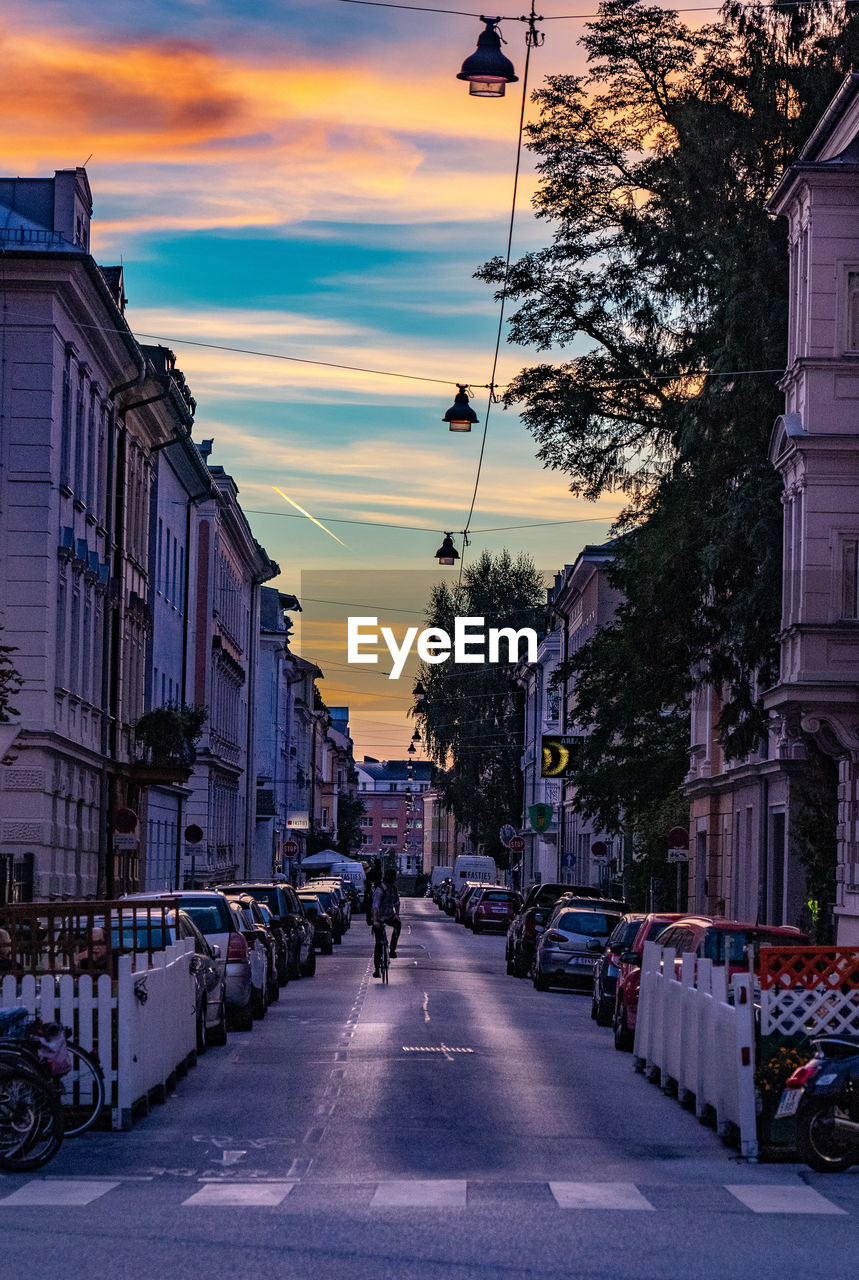 City street by buildings against sky during sunset