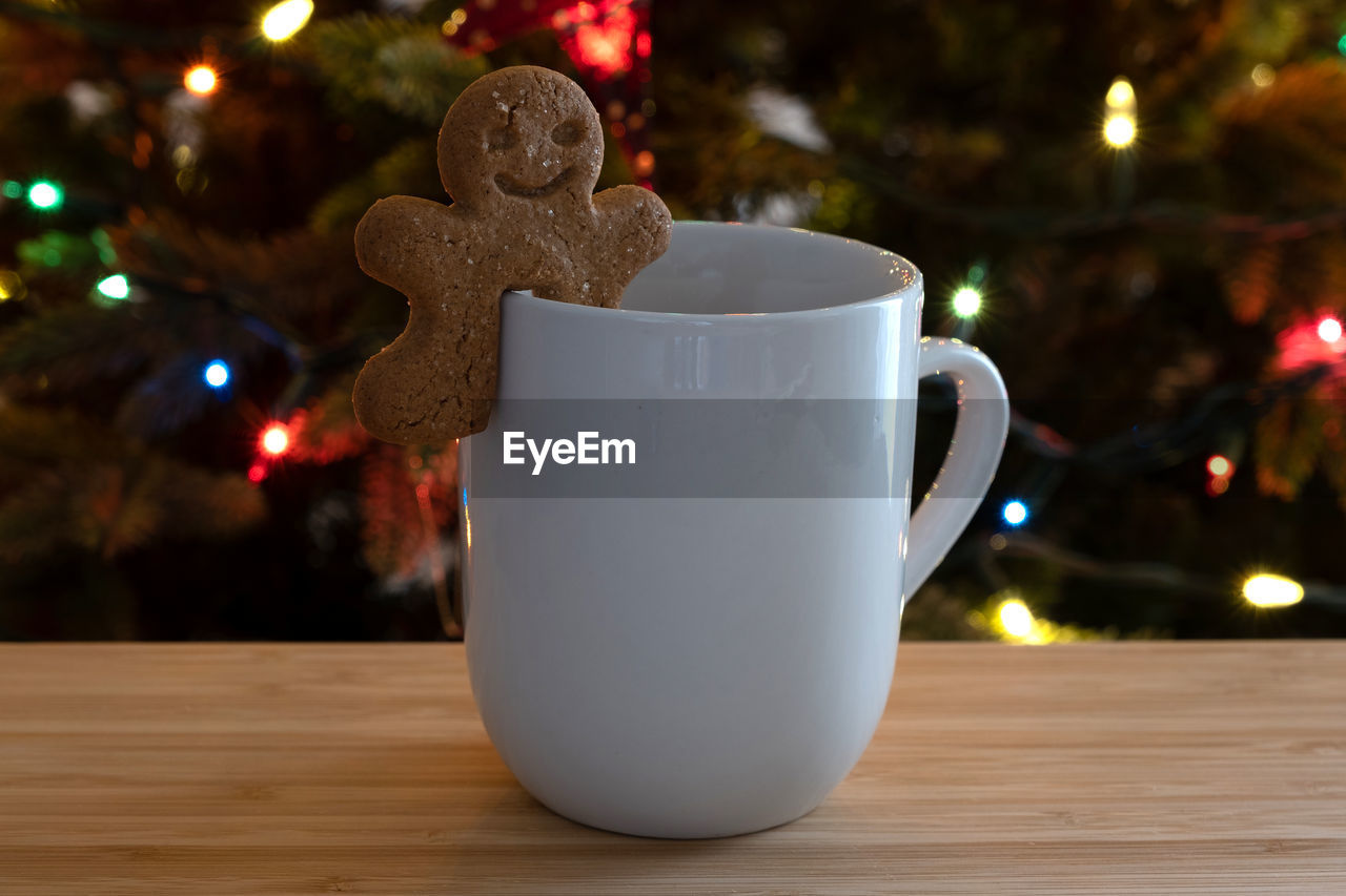 A holiday beverage in front of the christmas tree with a gingerbread man sitting on the rim 