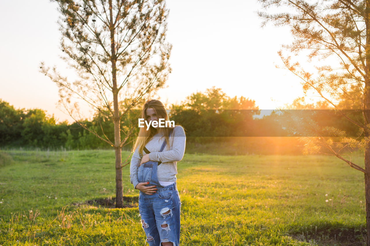 FULL LENGTH PORTRAIT OF WOMAN ON FIELD DURING SUNSET