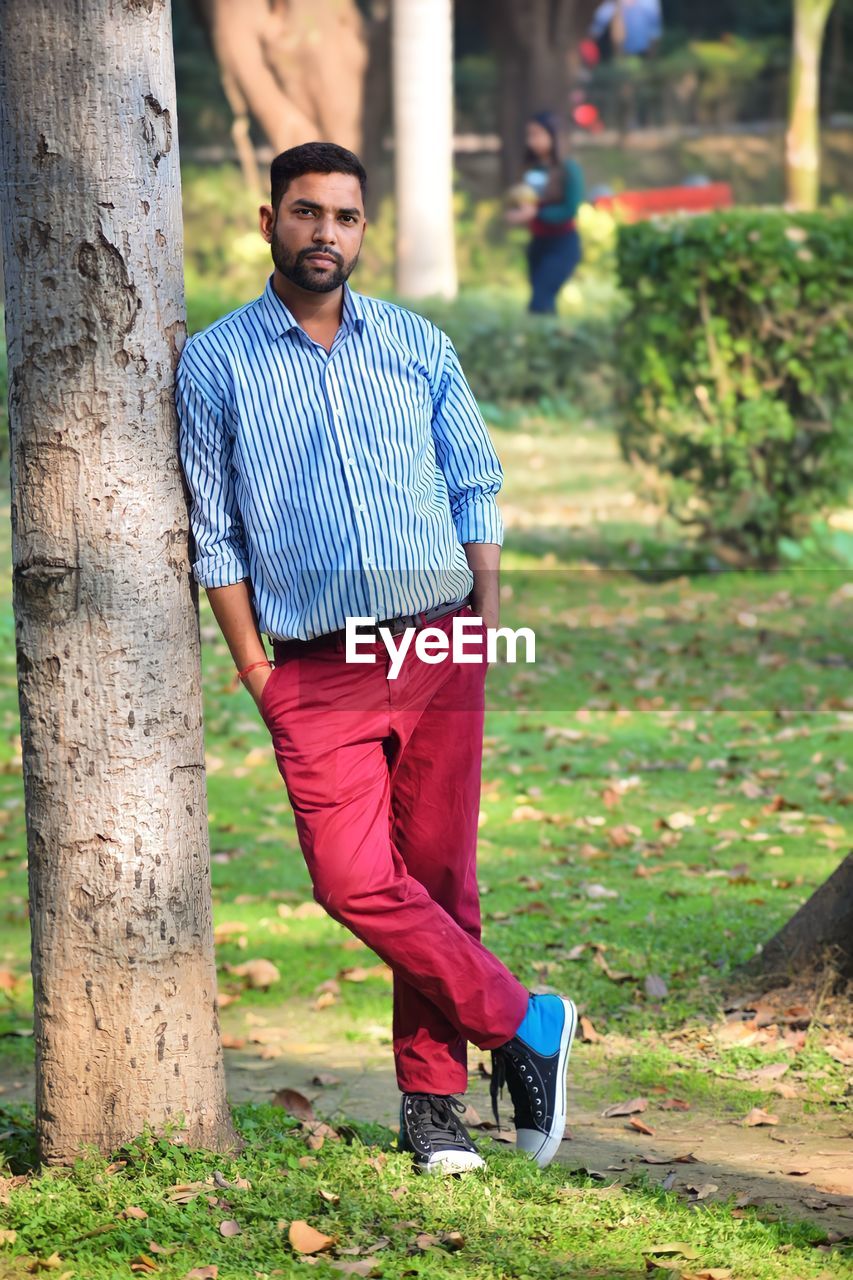 one person, spring, adult, men, green, tree, full length, lifestyles, casual clothing, portrait, plant, nature, young adult, standing, tree trunk, person, leisure activity, smiling, footwear, red, trunk, grass, relaxation, emotion, day, clothing, outdoors, happiness, sports, looking at camera, front view, beard, facial hair, autumn