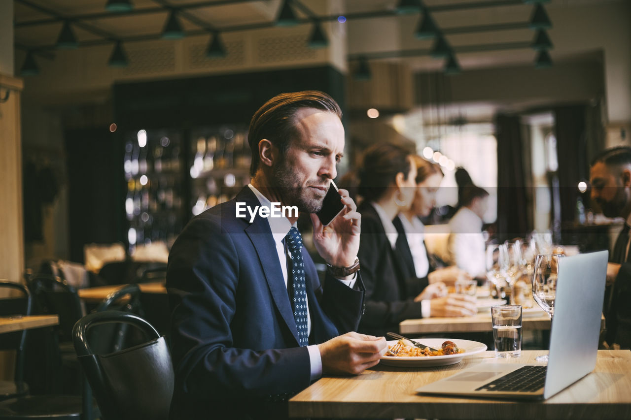 Male business person talking on phone while eating food in restaurant