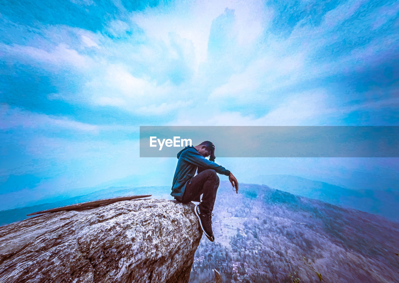 Side view full length of sad young man sitting on cliff against sky