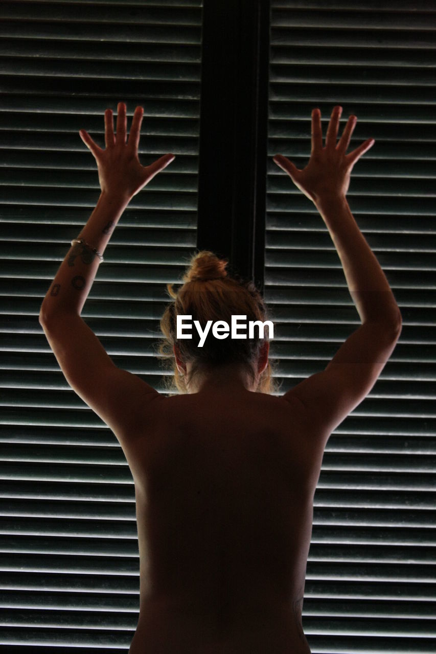 Rear view of naked woman with arms raised standing by window