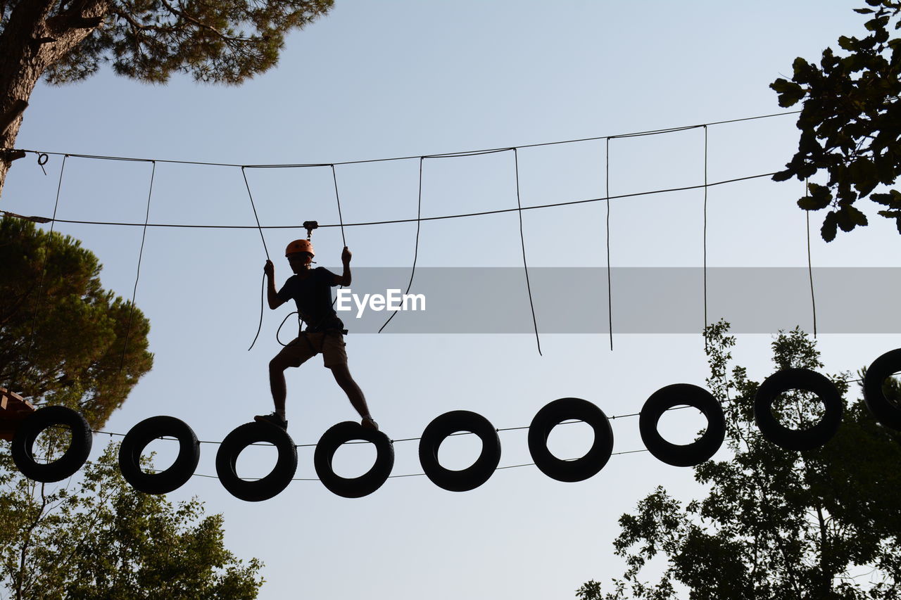 Low angle view of silhouette man on rope course against clear sky