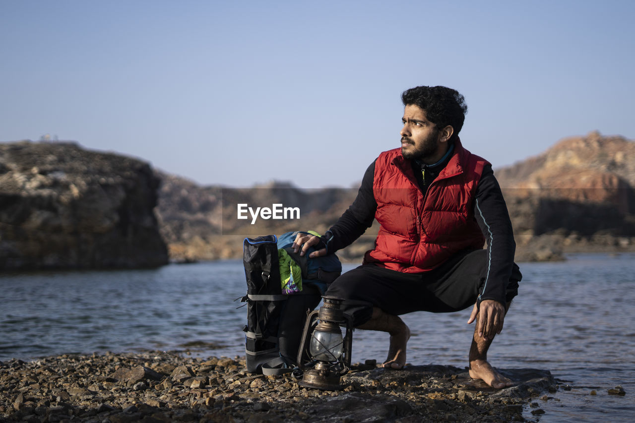 Young indian traveler getting ready for camping in the mountains, sitting near a lake with his pack.