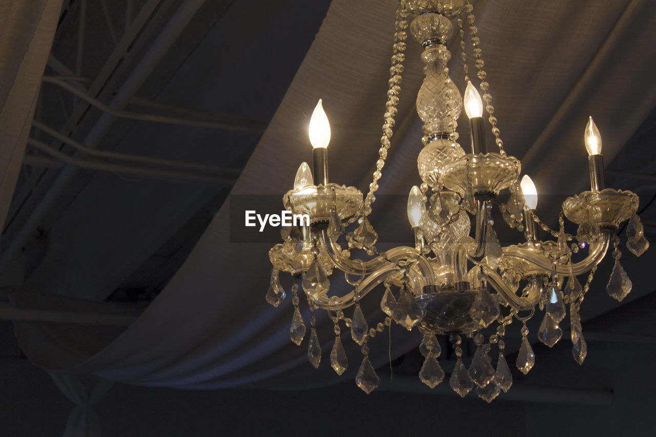 Low angle view of illuminated old chandelier hanging against fabric