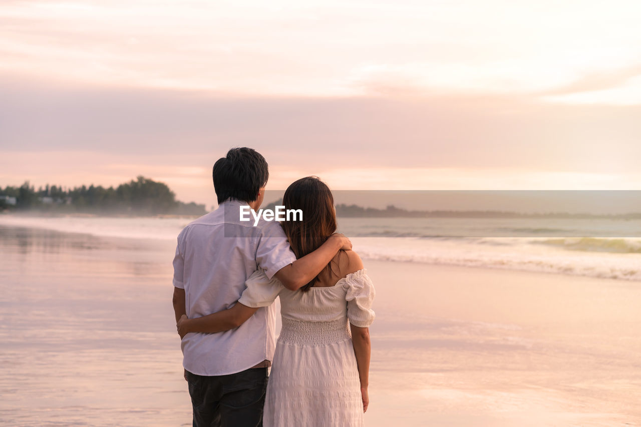 REAR VIEW OF COUPLE STANDING AT SEA AGAINST SKY