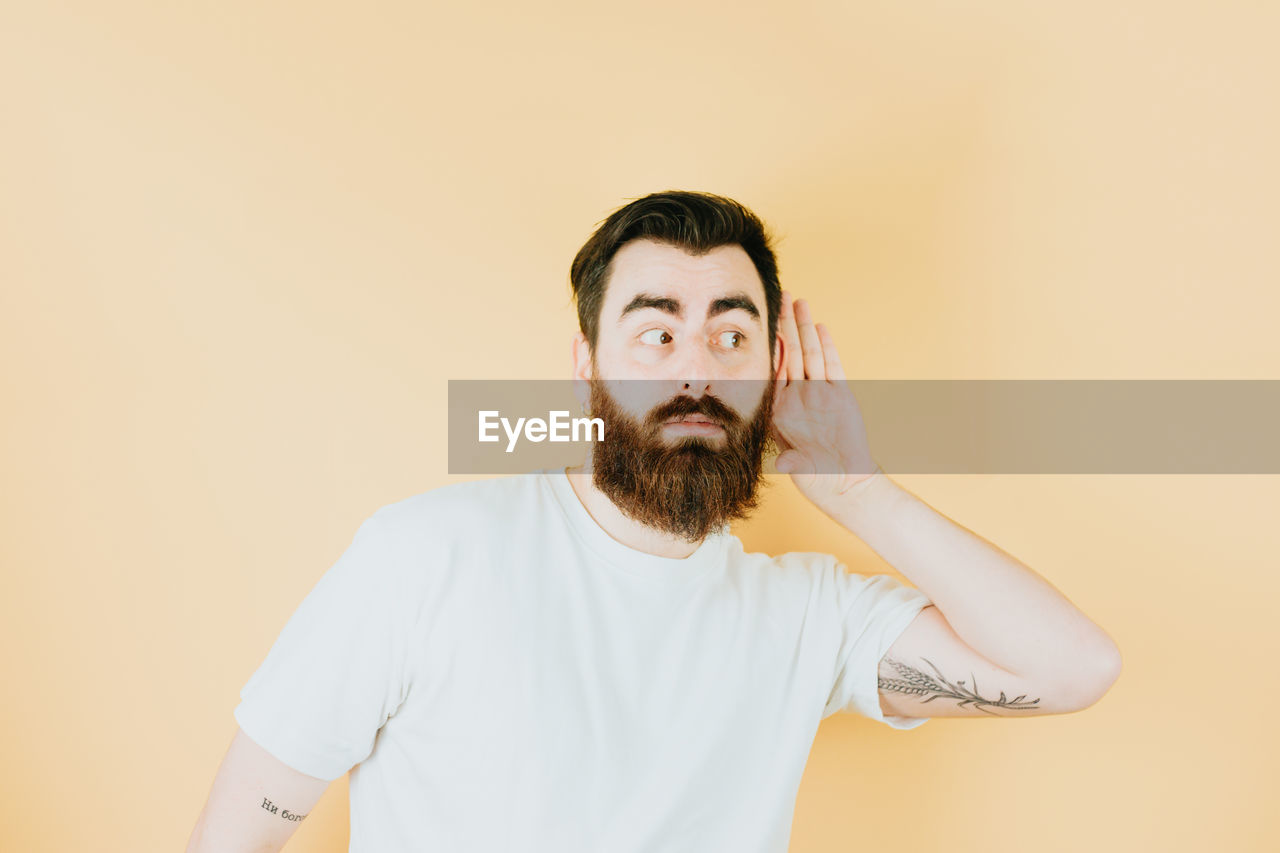 facial hair, beard, one person, adult, portrait, indoors, studio shot, men, waist up, colored background, copy space, moustache, person, human hair, front view, young adult, looking at camera, casual clothing, human face, standing, brown hair, looking, t-shirt, hairstyle, clothing, emotion, beige background, lifestyles, headshot