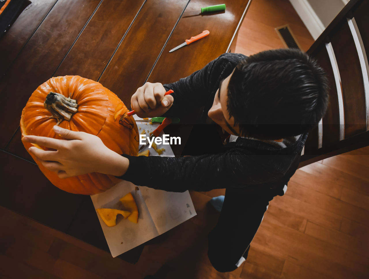 High angle view of boy making jack o' lantern of fresh pumpkin while sitting at table