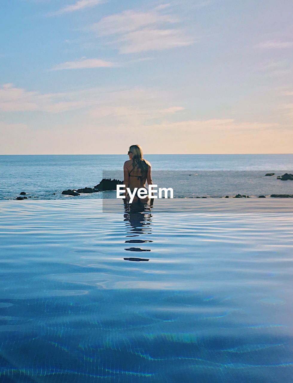 Rear view of woman swimming in infinity pool against sky during sunset