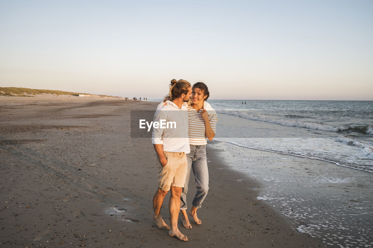 Young couple walking at beach against clear sky during vacation