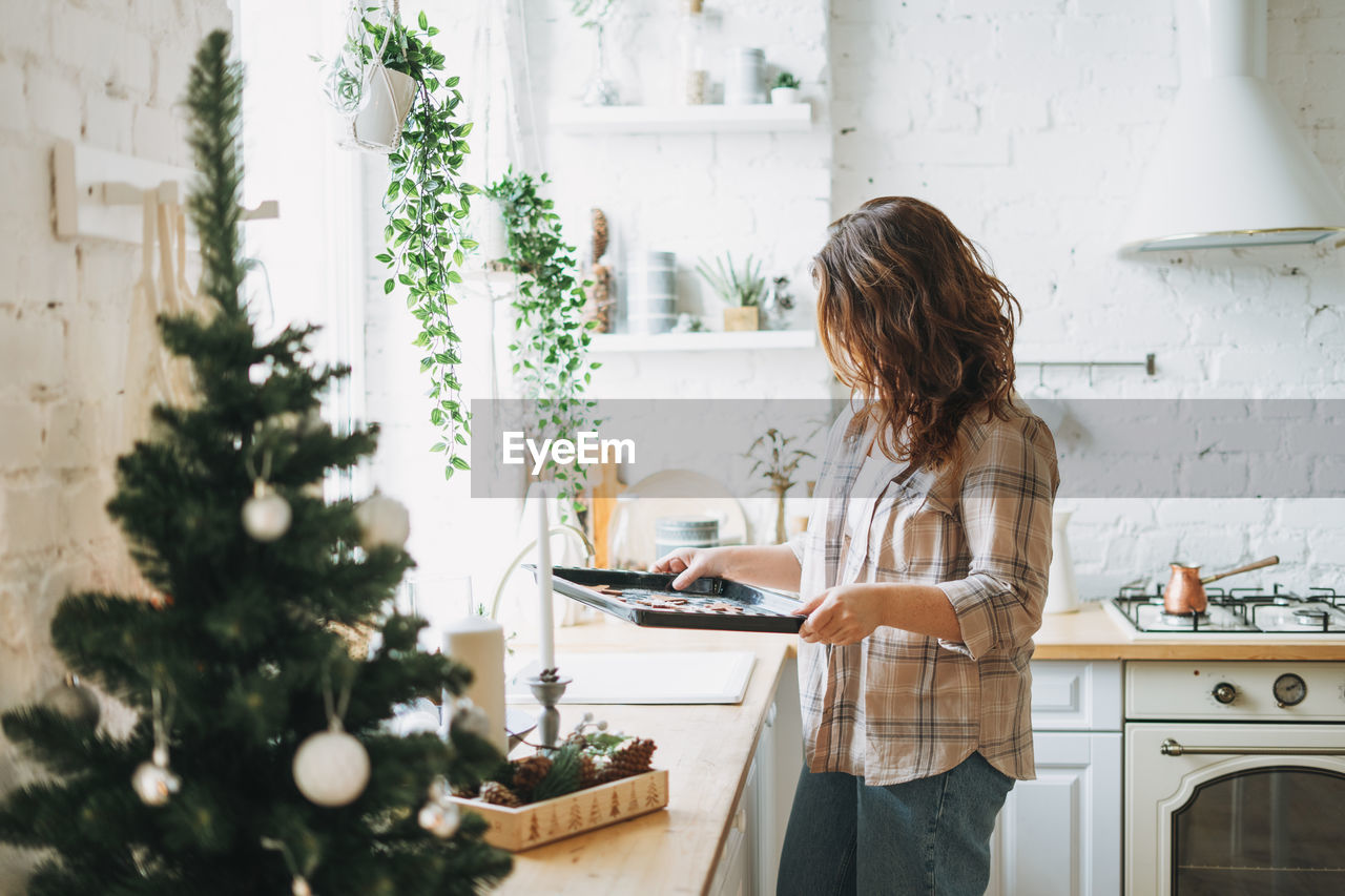 Attractive smiling woman with curly hair in plaid shirt bakes cookies at bright kitchen at home