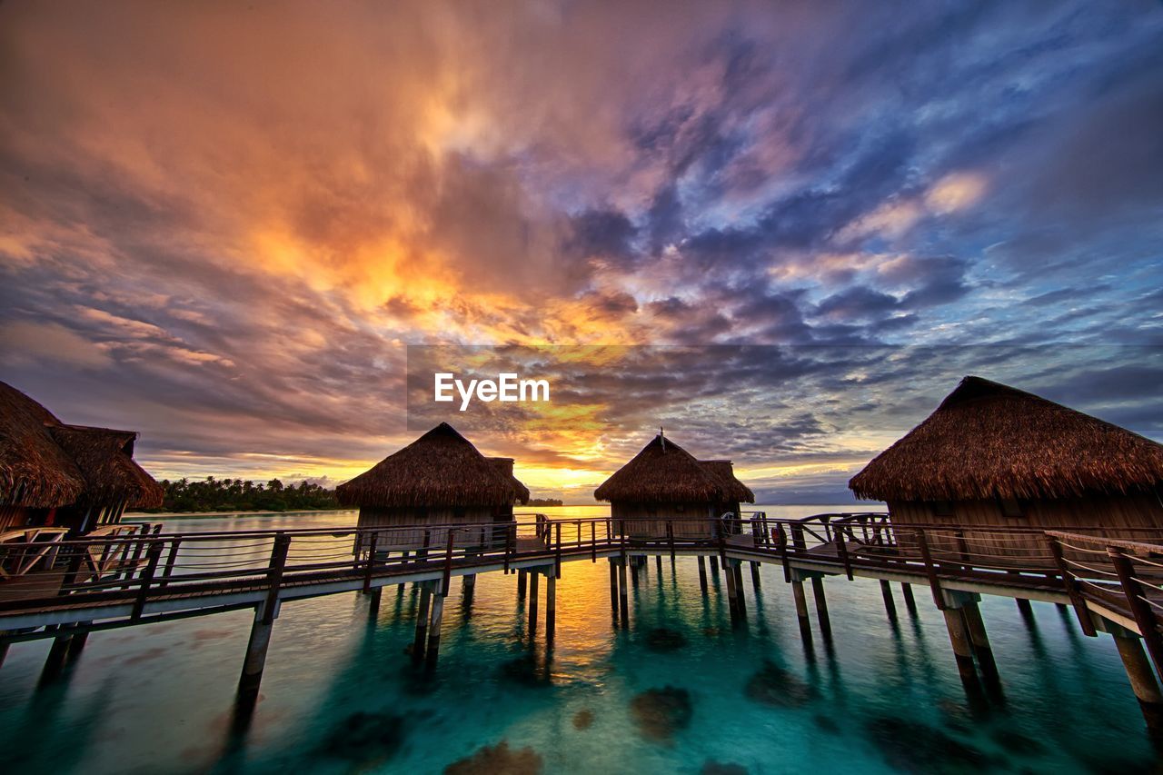 Stilt houses over sea against cloudy sky at sunset