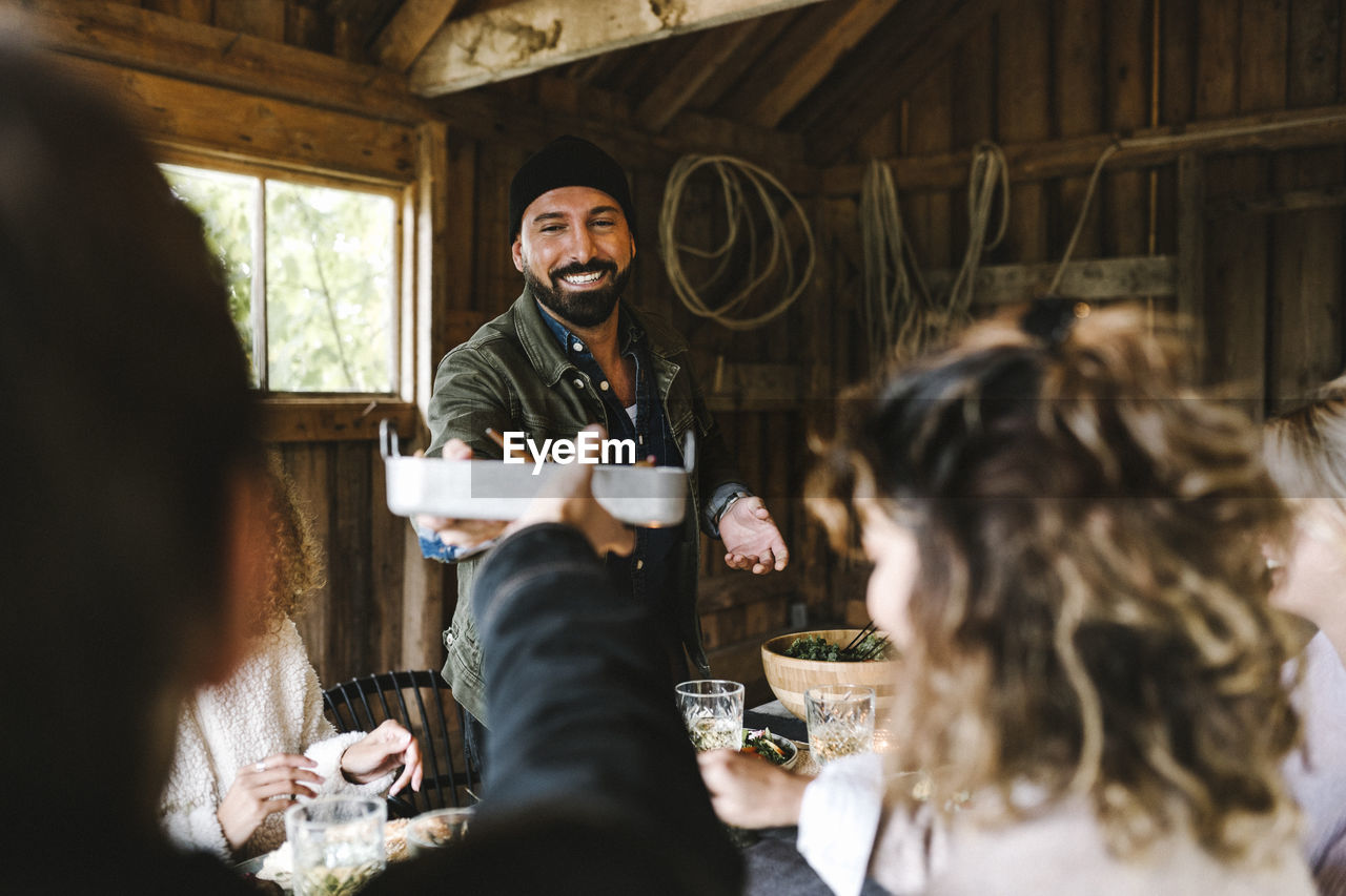 Smiling male passing food container to friend over dining table during social event