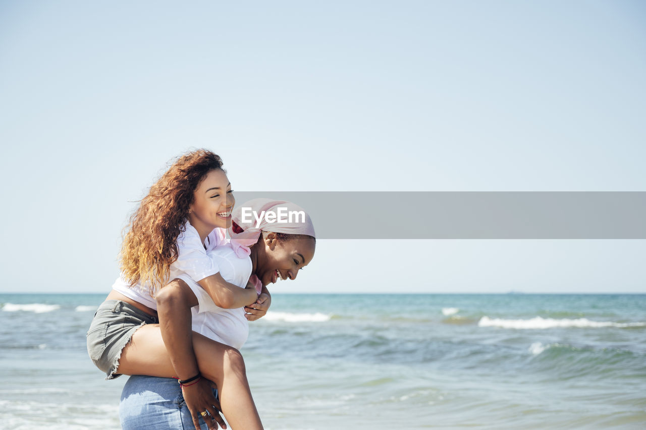 Delighted black mother piggybacking young daughter while having fun with closed eyes at seaside in summer