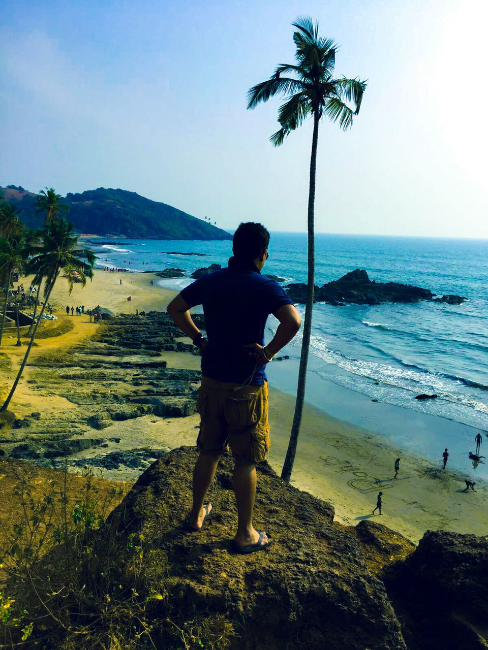 Full length rear view of man with hands on hip standing on rock formation at vagator beach