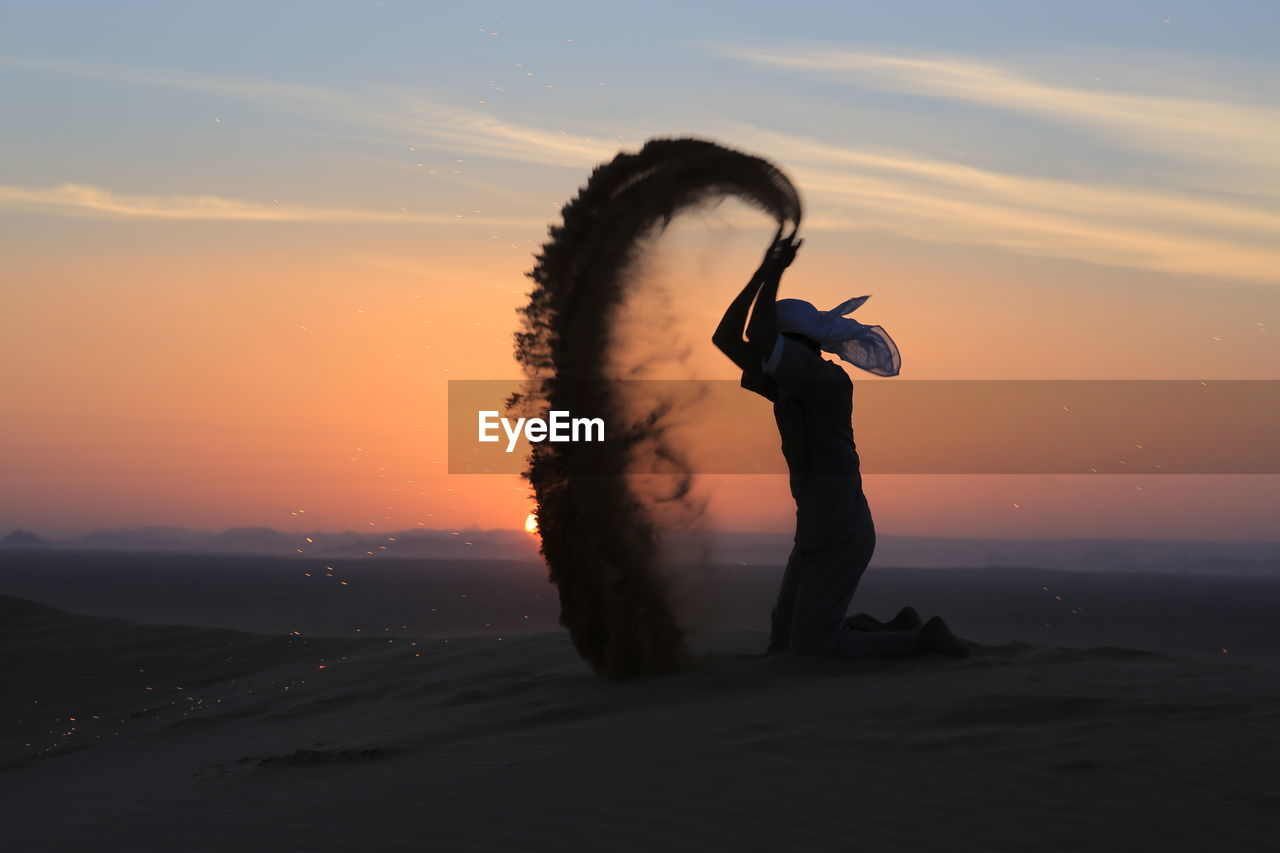 Silhouette man throwing sand at desert against sky during sunset