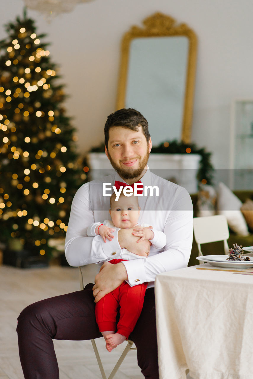 A stylish dad in a white shirt with a red bow tie holds his son in his arms against the background