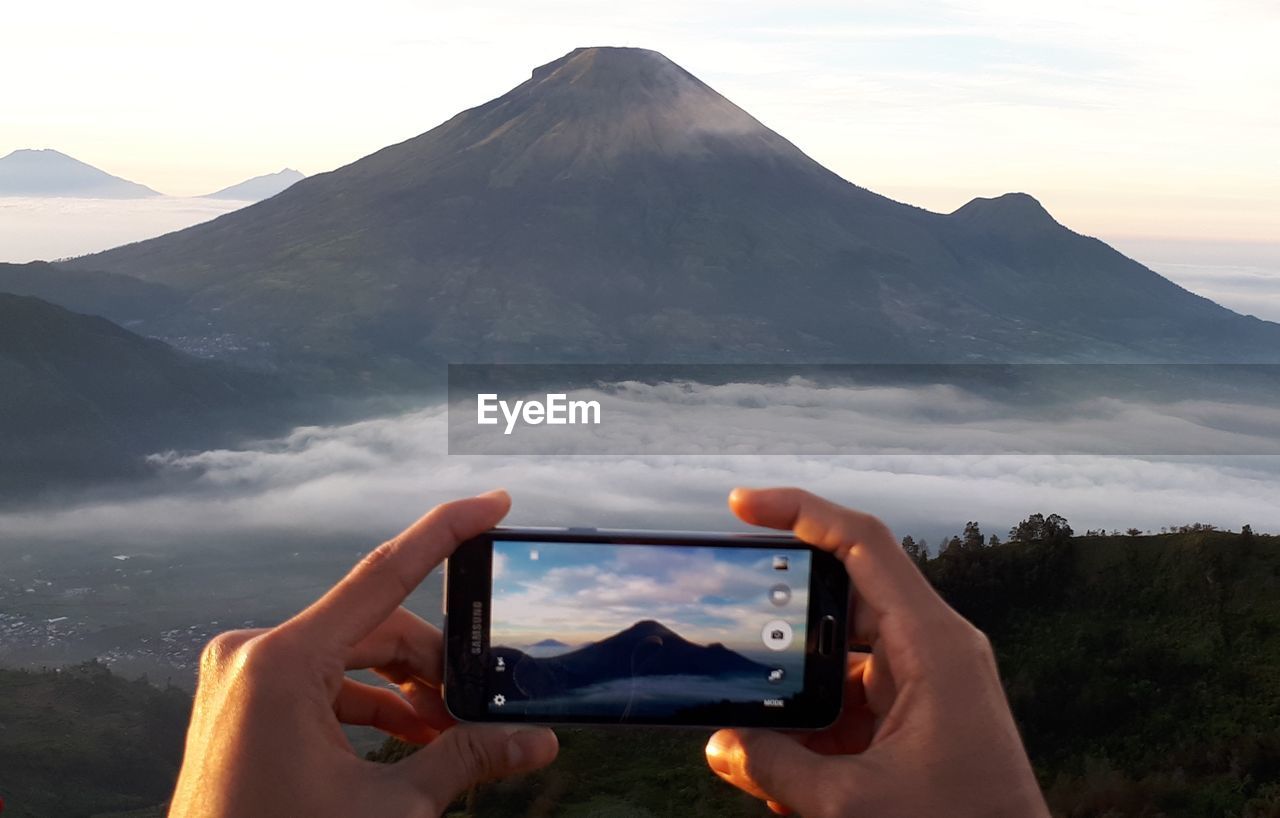 MIDSECTION OF PERSON PHOTOGRAPHING MOBILE PHONE AGAINST MOUNTAIN