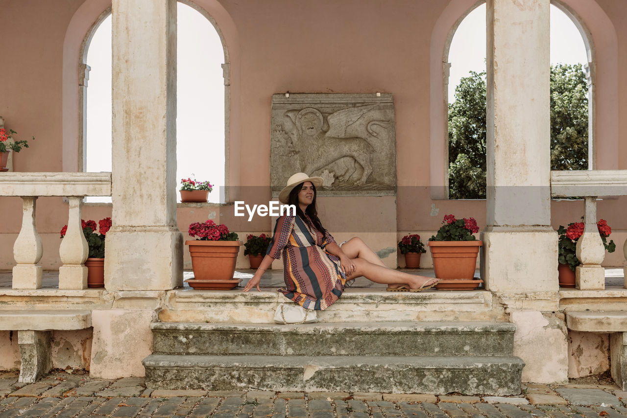 Lifestyle photo of stylish young woman sitting on stone stairs under arched columns of old building