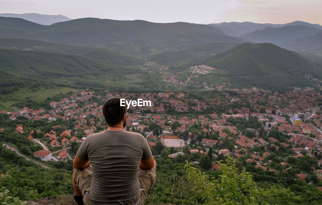 Rear view of man looking at town while sitting on mountain