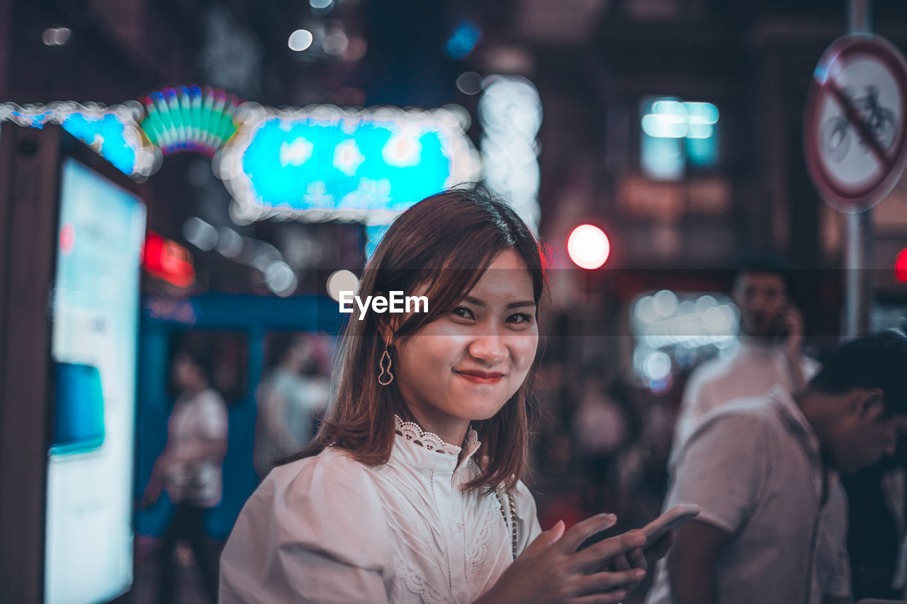 PORTRAIT OF SMILING YOUNG WOMAN STANDING IN CITY AT NIGHT