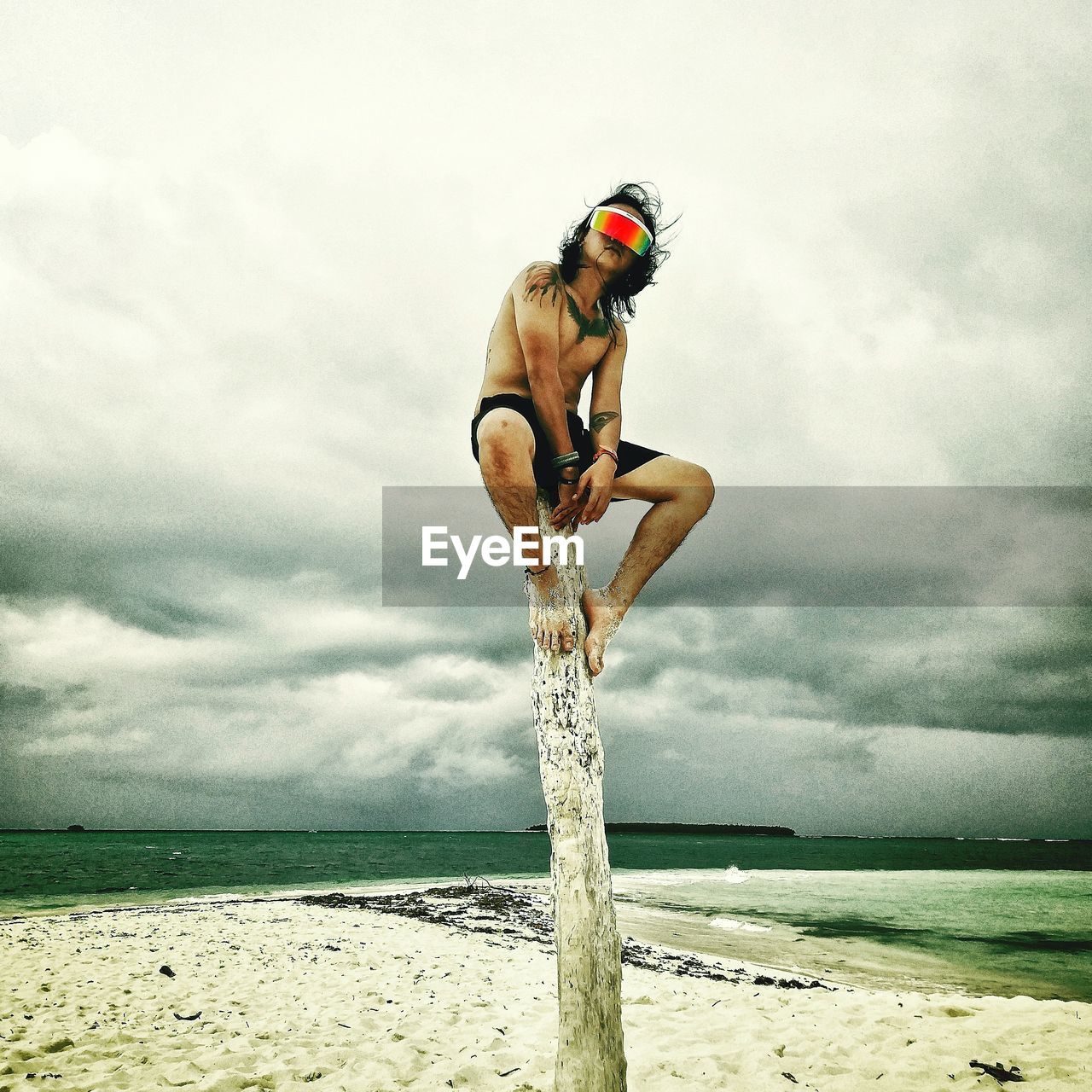 Man wearing a virtual reality headset while sitting on wood at beach against cloudy sky