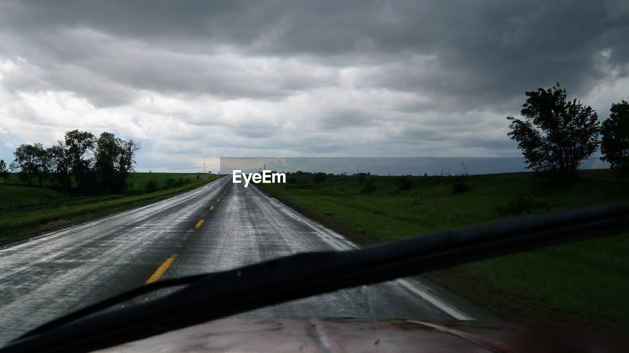 Road amidst grassy field against cloudy sky seen through car windshield